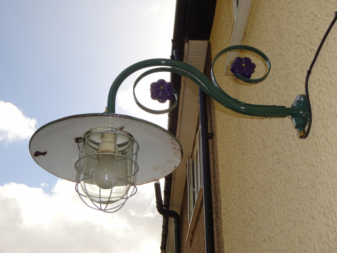 Simplex enamel wellglass light and Revo B28 bracket installed.
It only took me 5 years, but yesterday the massive Simplex well glass light I restored, and then had to strip and restore again, has finally been installed as our permanent motion sensor light, and I absolutely love the way it turned out.

When I originally bought this light, it was an absolute mess, missing the glass, bent, buckled and rusted cage, missing screws, and a bracket that consisted of a conduit box and angle metal, it took a lot of work to rebuild it back to a complete state once again, even going to the effort of having a glass replicated. 

The Revo B28 bracket came from eBay, and upon arrival I realised it had the correct conduit reduction adapter fitted to allow me to install this massive light to. After stripping the paint from the enamel shade revealing the original green, I clear lacquered it, and then had it colour matched so I could paint the bracket in the matching colour, painting the flowers purple for a little contrast.

Installation was actually remarkably easy, it only took about half an hour to get it on the wall and wired up. This was the first time I saw the light actually lit in the time I've had it, and with an Omega 150w GLS lamp, it looks absolutely stunning. It provides ample light for the patio area its intended for, and the motion sensor switches on perfectly where I want it to as well. I've also wired in a manual override too, which will come in mega handy for when the lights needed longer than the time set on the PIR.
