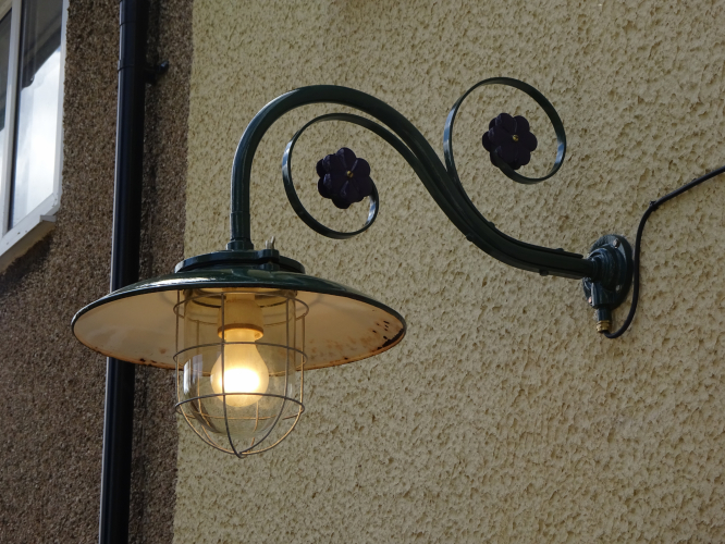 Simplex enamel wellglass light and Revo B28 installed and lit.
It only took me 5 years, but yesterday the massive Simplex well glass light I restored, and then had to strip and restore again, has finally been installed as our permanent motion sensor light, and I absolutely love the way it turned out.

When I originally bought this light, it was an absolute mess, missing the glass, bent, buckled and rusted cage, missing screws, and a bracket that consisted of a conduit box and angle metal, it took a lot of work to rebuild it back to a complete state once again, even going to the effort of having a glass replicated. 

The Revo B28 bracket came from eBay, and upon arrival I realised it had the correct conduit reduction adapter fitted to allow me to install this massive light to. After stripping the paint from the enamel shade revealing the original green, I clear lacquered it, and then had it colour matched so I could paint the bracket in the matching colour, painting the flowers purple for a little contrast.

Installation was actually remarkably easy, it only took about half an hour to get it on the wall and wired up. This was the first time I saw the light actually lit in the time I've had it, and with an Omega 150w GLS lamp, it looks absolutely stunning. It provides ample light for the patio area its intended for, and the motion sensor switches on perfectly where I want it to as well. I've also wired in a manual override too, which will come in mega handy for when the lights needed longer than the time set on the PIR.
